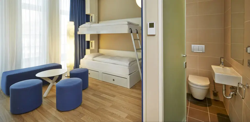 Sitzgruppe aus blauen Hockern und einem kleinen Tisch vor einem Etagenbett. Auf der rechten Seite steht die Tür offen und man sieht eine Toilette und ein Waschbecken.