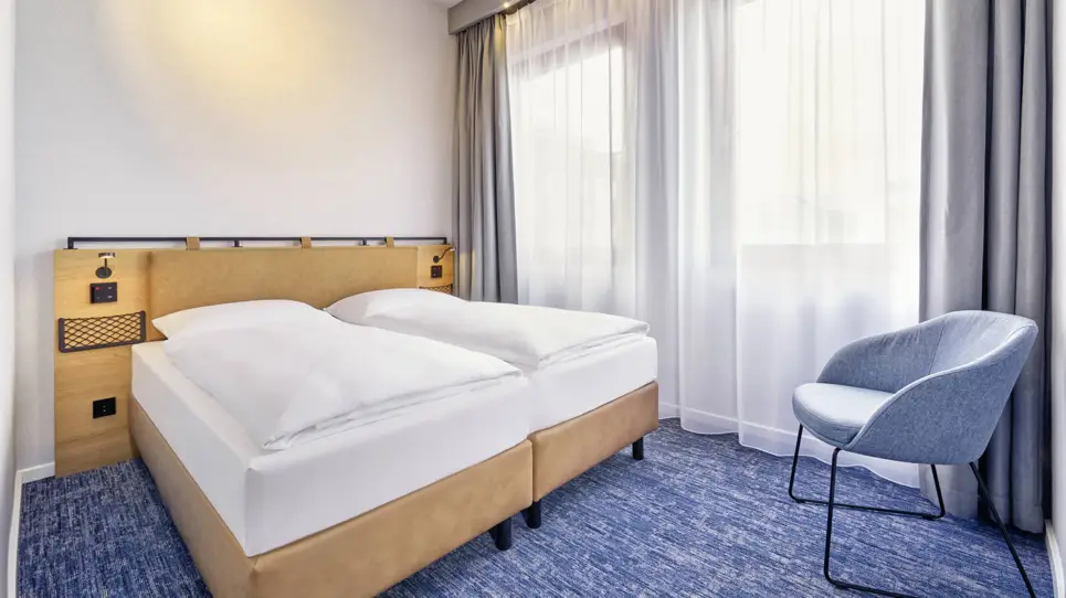 Doppelbett mit kleinen Taschen an der Rückwand. In der Ecke steht ein Stuhl.