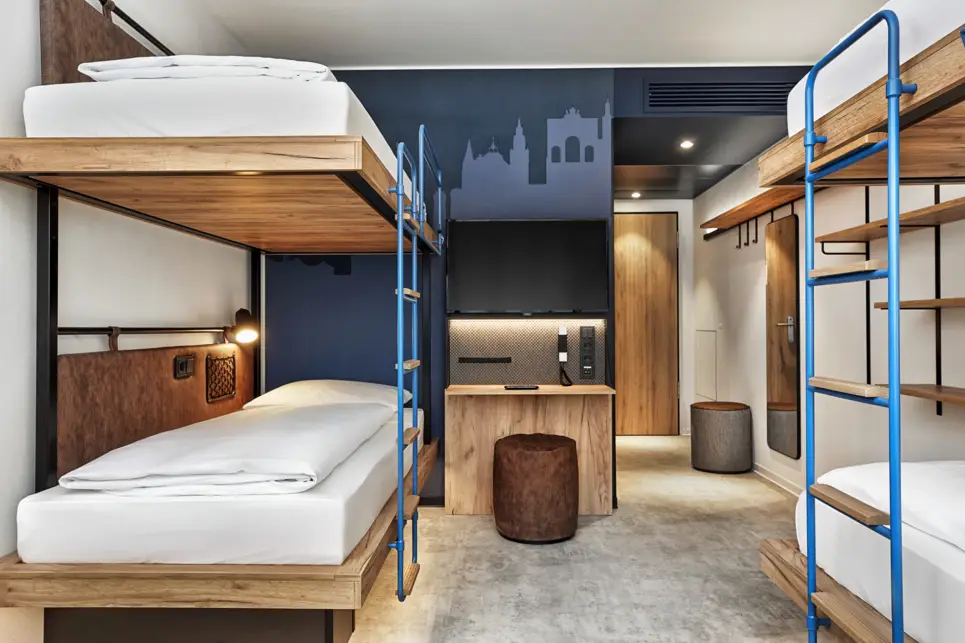 Vierbettzimmer mit Etagenbetten in dunkelbraunem Holz und Leder. An der Wand ist die Münchener Skyline in verschiedenen Blautönen gemalt.