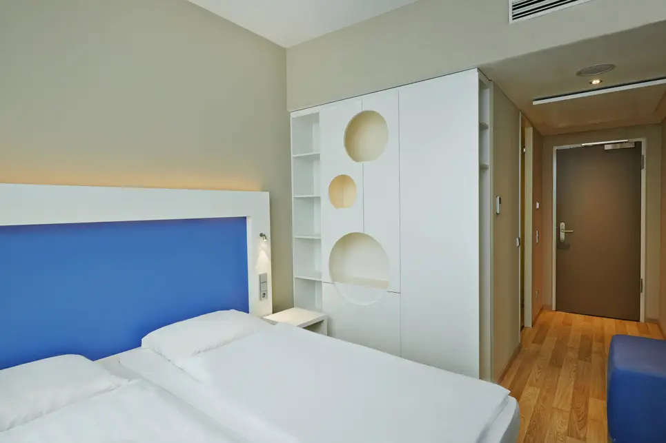 Doppelbett mit einer blauen Rückwand. Neben dem Bett steht ein großer, weißer Kleiderschrank.