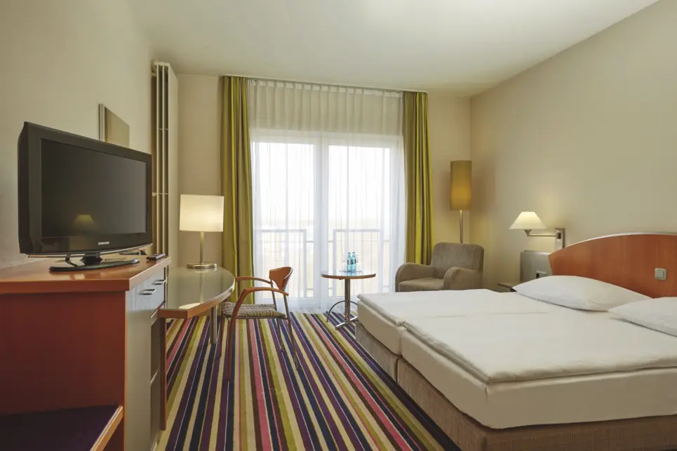 Hotelzimmer mit einem Doppelbett rechts und einem Fernseher links. Neben dem Fernsheer stet ein großer Schreibtisch.