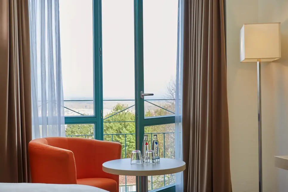 Eine Sessel und ein kleiner Tisch stehen vor einem geöffneten Fenster im Hotelzimmer.