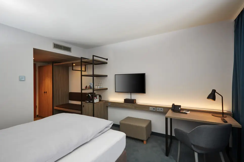 Blick in das Zimmer mit einem Einzelbett. Vor dem Bett steht ein kleiner Schrank, ein Schreibtisch und an der Wand hängt ein Fernseher.