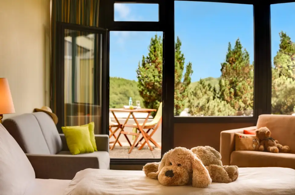 Nahaufnahme auf das Bett mit einem Kuscheltierhund darauf. Im Hintergrund ist der Balkon mit dem Blick auf einen Wald.