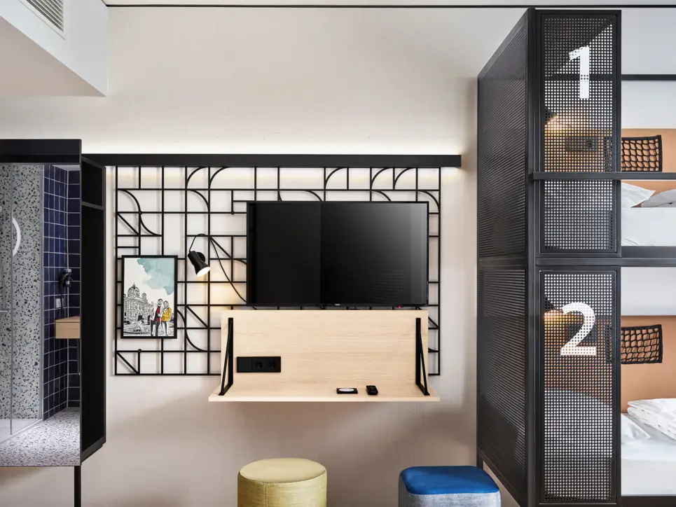 Schreibtisch mit einem Fernseher und Hockern. Daneben stehen Etagenbetten und eine kleine Gaderobe.