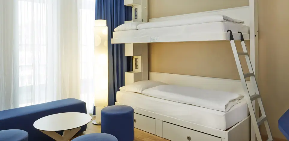 Etagenbett vor einem Fenster. Vor dem Bett steht eine kleine Sitzgruppe mit einem Tisch.