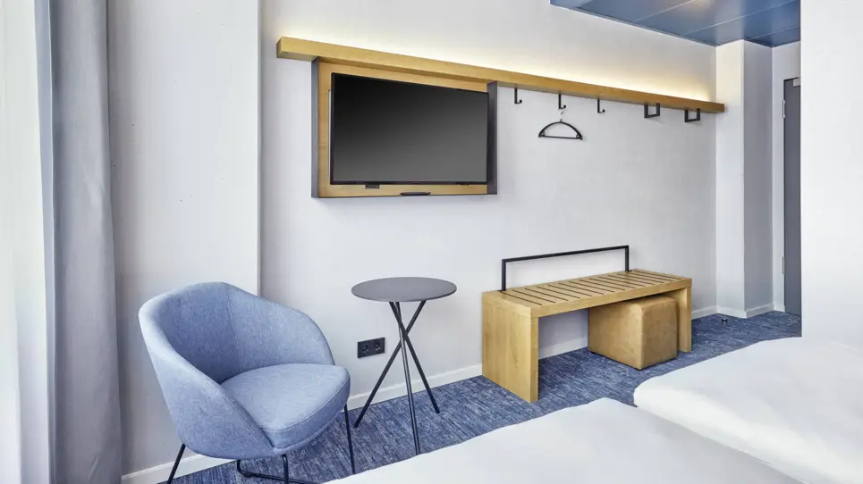 Gegenüber vom Bett steht ein Stuhl mit einem kleinen Tisch und einer Ablagebank. An der Wand hängt ein Fernseher.