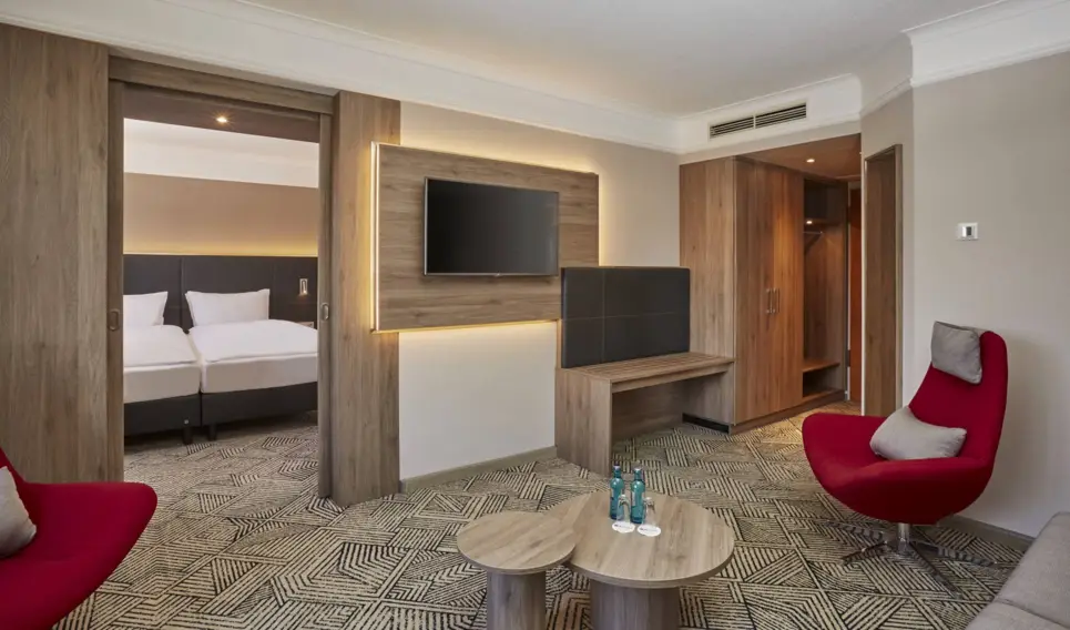 Wohnbereich der Suite mit einem Sessel, einem Fernseher und ein Tisch. Auf der linken Seite ist eine Tür mit Blick auf das Doppelbett.