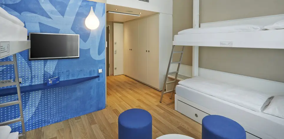 Etagenbett im Hotelzimmer. Vor dem Bett stehen zwei blaue Hocker.