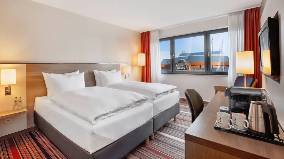 Ein Schreibtisch mit einer Kaffeemaschine vor einem Doppelbett. Aus dem Fenster sieht man das Musical-Theater.