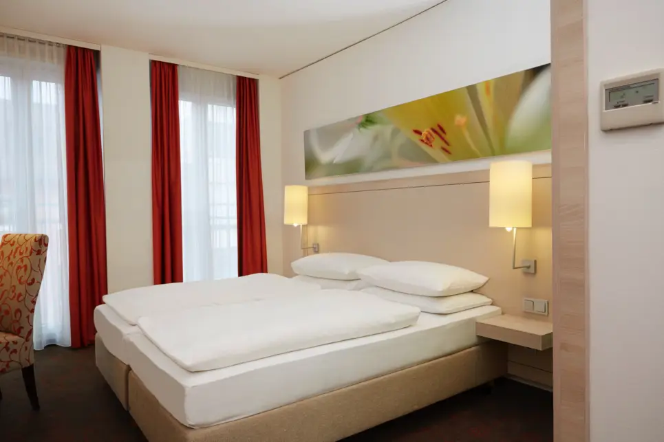 Großes Doppelbett im Hotelzimmer. Am Bett hängen zwei kleine Lampen.