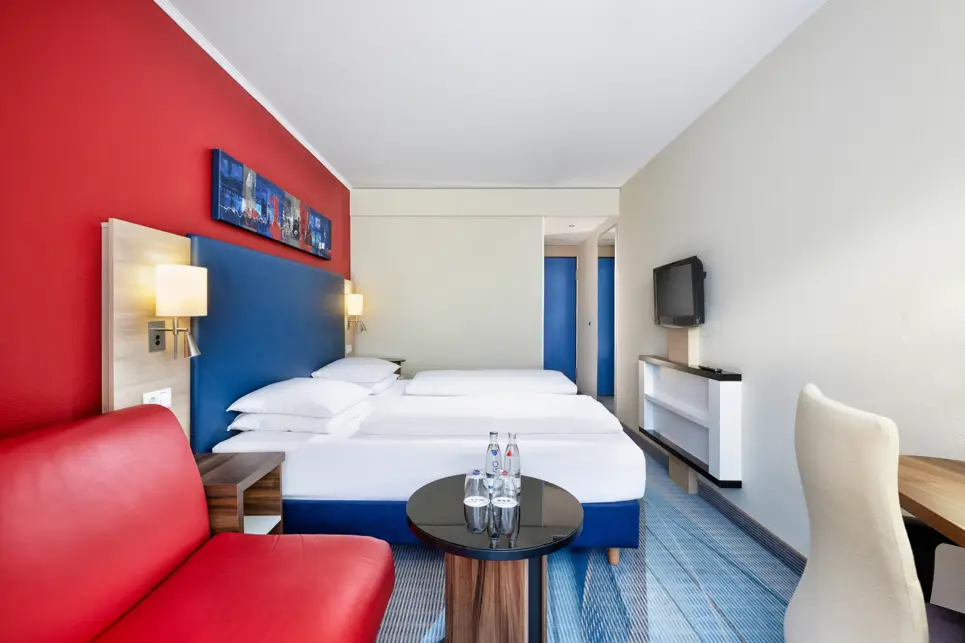 Hotelzimmer mit einem Doppelbett und einem Fernseher an der Wand. Neben dem Bett steht ein Nachttisch.