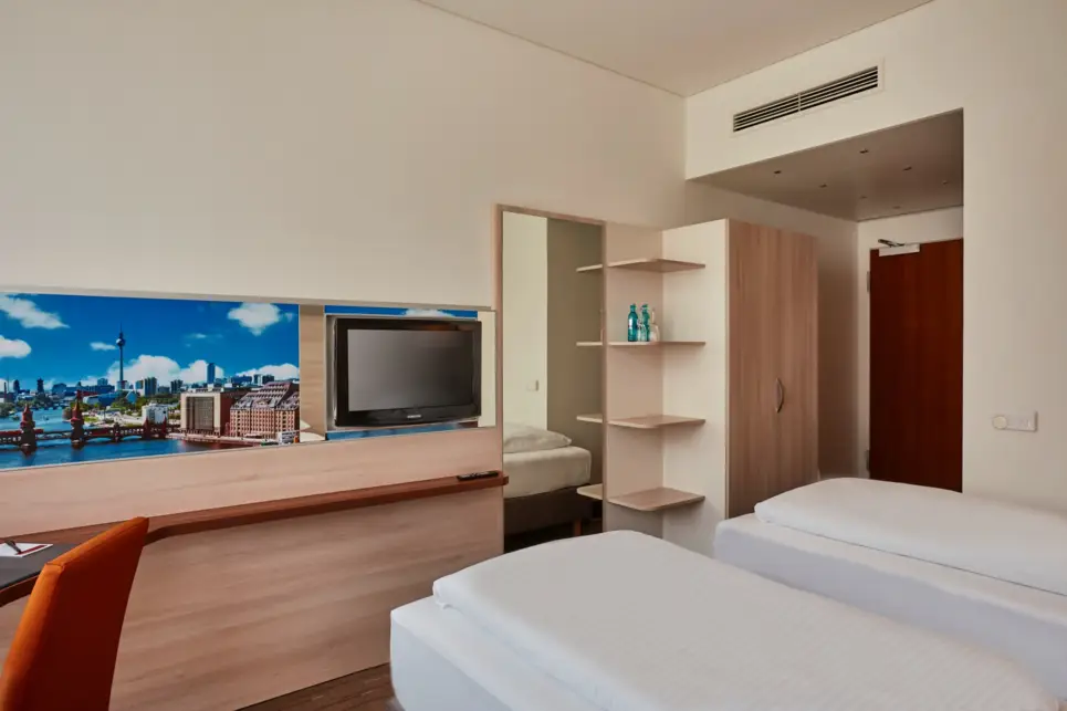 Twinbetten im Hotelzimmer. Am Fußende hängt ein Fernseher und ein großer Spiegel.