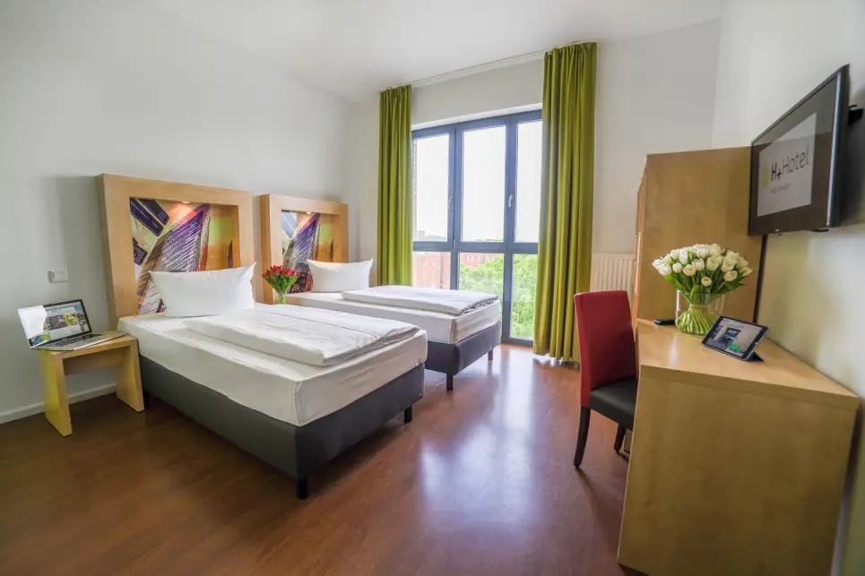 Zwei Einzelbetten vor einem großen Fenster. Vor den Betten steht ein Schreibtisch mit einem Stuhl und ein Kleiderschrank.