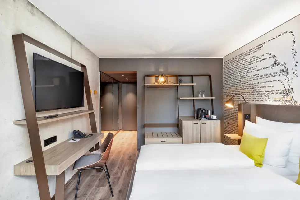 Doppelbett mit einem kleinen Tisch und Stuhl steht vor einem Regal mit Schrank. Über dem Tisch hängt ein Fernseher an der Wand.