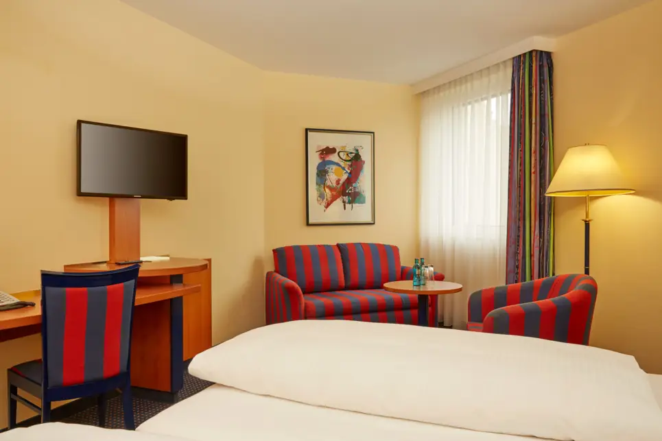 Hotelzimmer mit einer kleinen Sitzecke. Vor dem Bett ist ein kleiner Schreibtisch mit einem Fernseher.