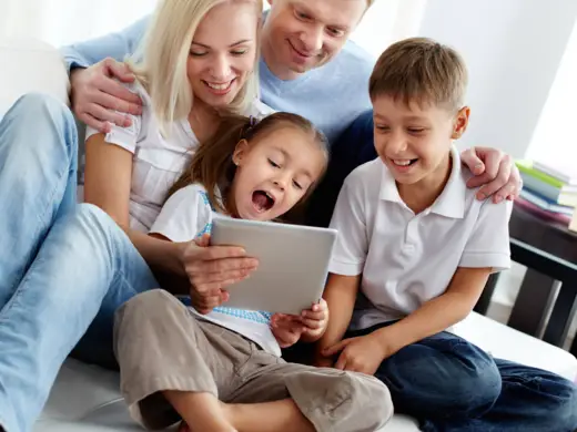 Eine vierköpfige Familie sitzt auf einer Couch und schaut freudig auf ein Tablet.