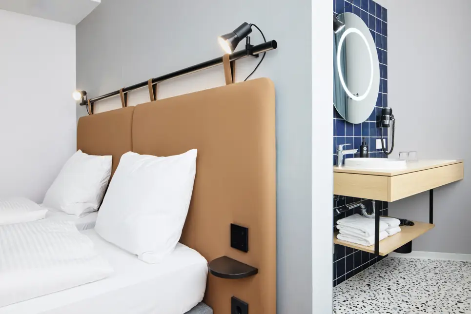 Nahaufnahme vom Bett. Hinter der Trennwand am Bett ist ein offenens Badezimmer.