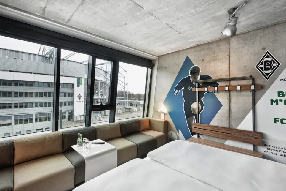 Hotelzimmer mit Blick auf das Stadion. Am Fenster steht eine graubraune Couch mit einem kleinen Tisch.