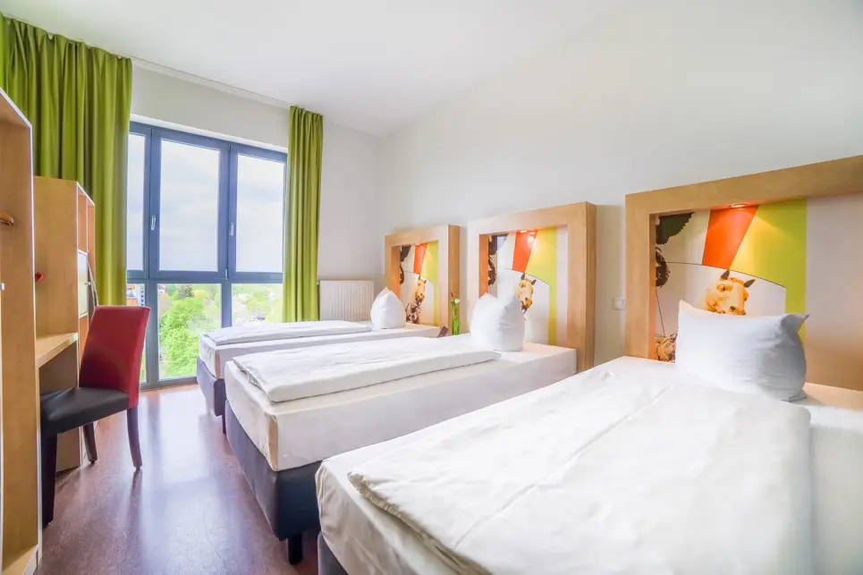 Drei Einzelbetten nebeneinander im Hotelzimmer. Vor den Betten steht ein Schreibtisch mit einem Stuhl.