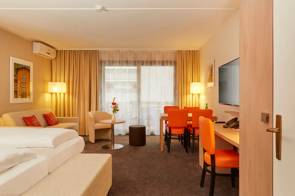 Hotelzimmer mit einem Doppelbett und einer Sitzecke. Auf der rechten Seite ein kleiner Esstisch und ein Schriebtisch.