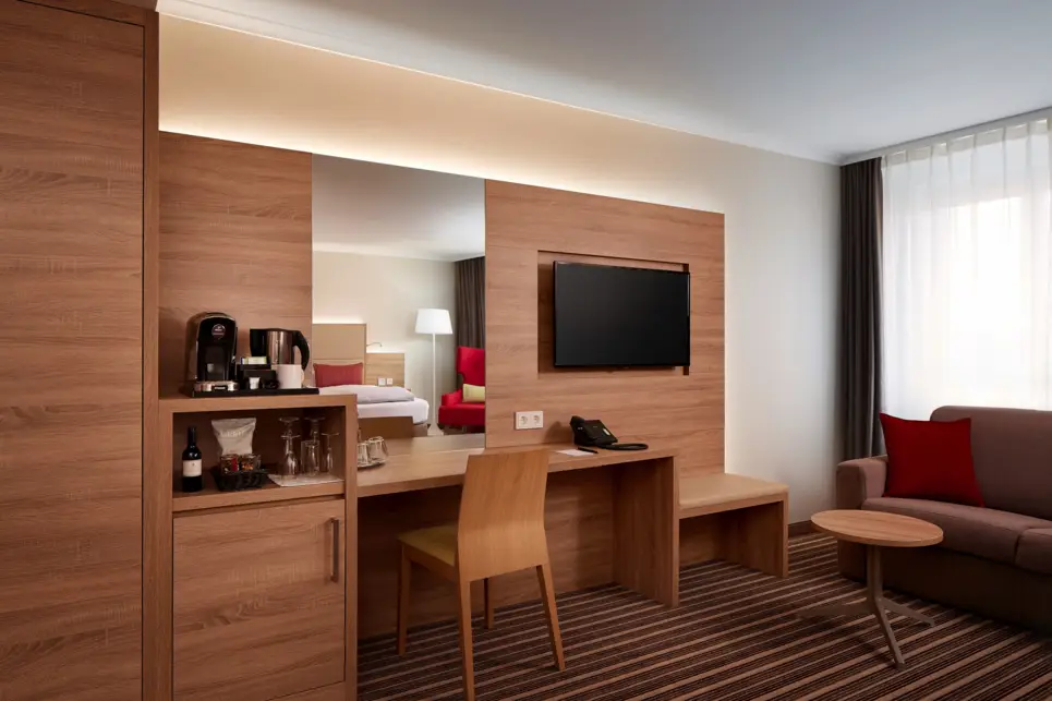 Holzverkleidete Wand mit indirekter Beleuchtung. Davor steht ein Schreibtisch und eine Minibar mit einer Kaffeemaschine.