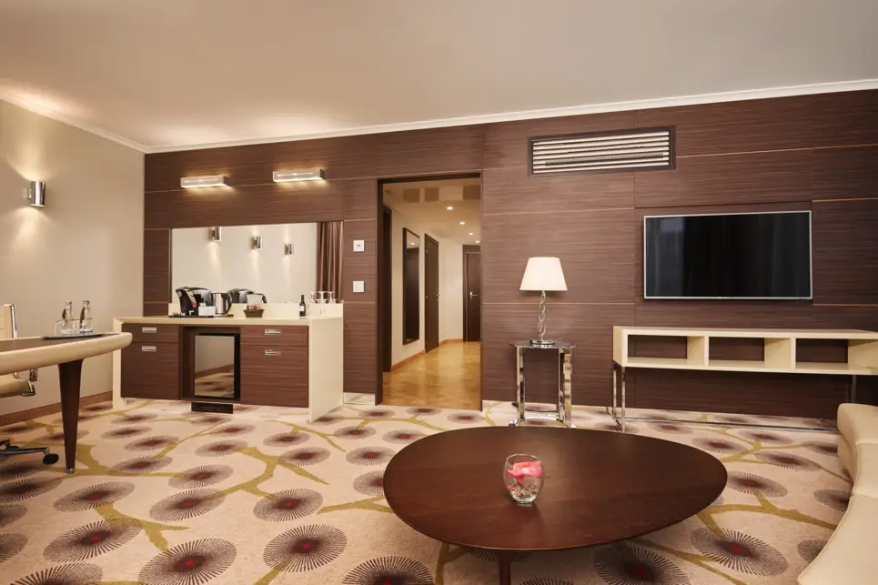 An der mit braunem Holz verkleideten Wand steht ein heller Schreibtisch links und hängt ein Fernseher rechts. Im Vordergrund steht ein großer, brauner Couchtisch.