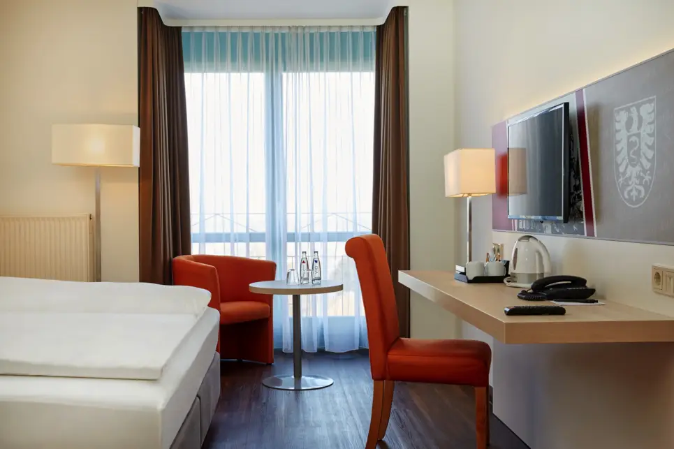 Schreibtisch vor dem Bett. Am Fenster steht ein Sessel.