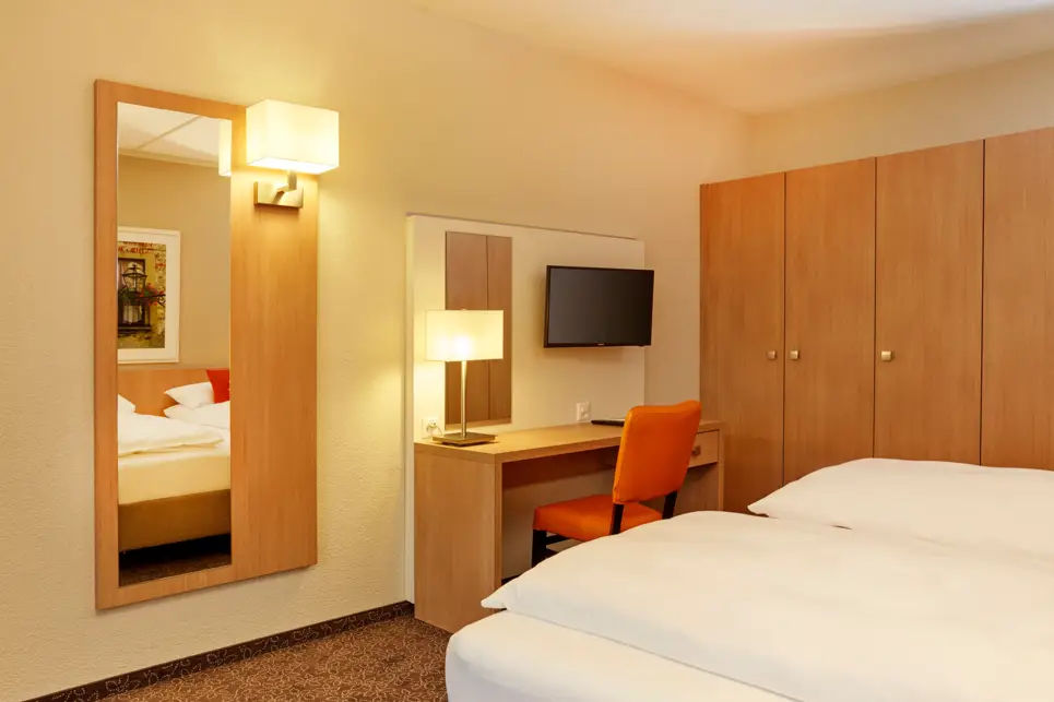 Hotelzimmer mit einem Doppelbett und einem Schreibtisch davor. Neben dem Schreibtisch ist ein großer Spiegel und ein großer Schrank.