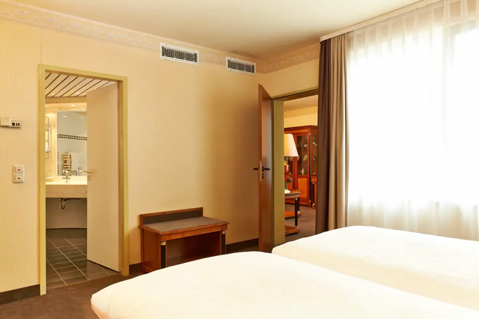 Schlafzimmer in der Suite mit einem Doppelbett und einer kleinen Ablage. Durch eine Tür ist das Badezimmer und in der anderen der Blick in den Wohnbereich.